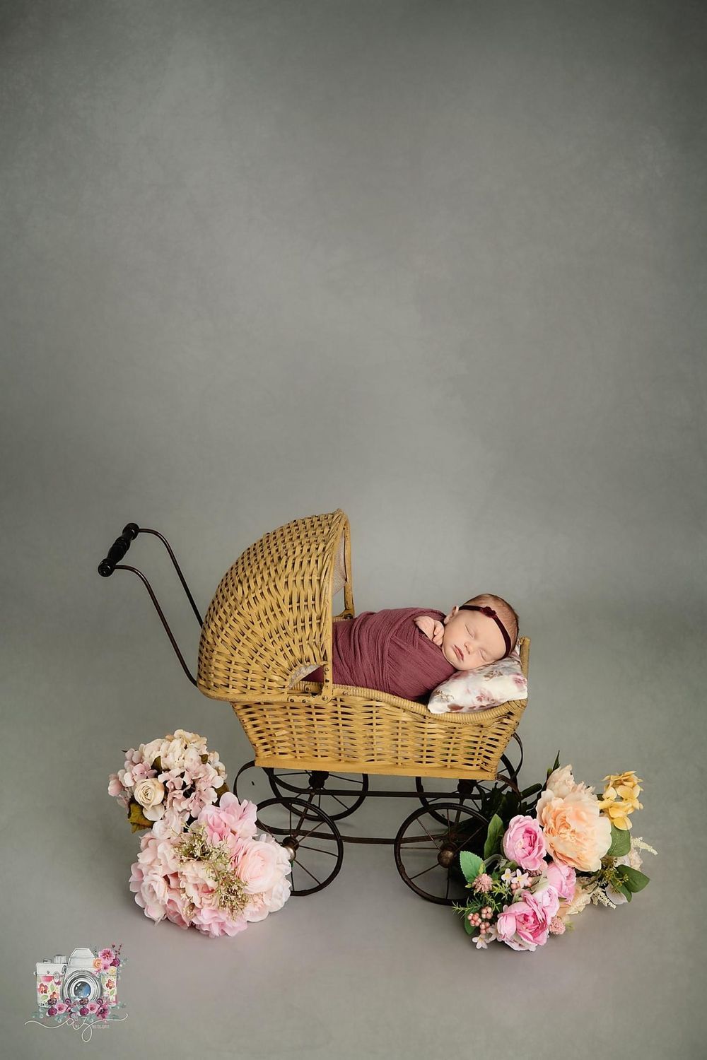 Vintage wicker baby carriage with sleeping infant surrounded by pink flowers against grey background.
