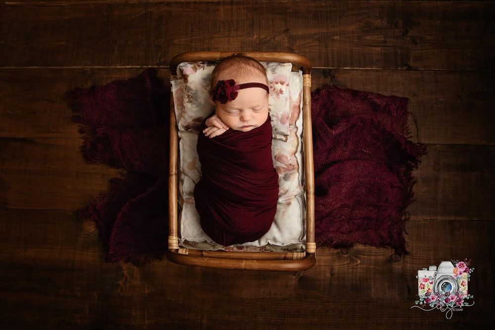 Sleeping newborn in burgundy wrap lying in rustic wooden box with textured fabric.