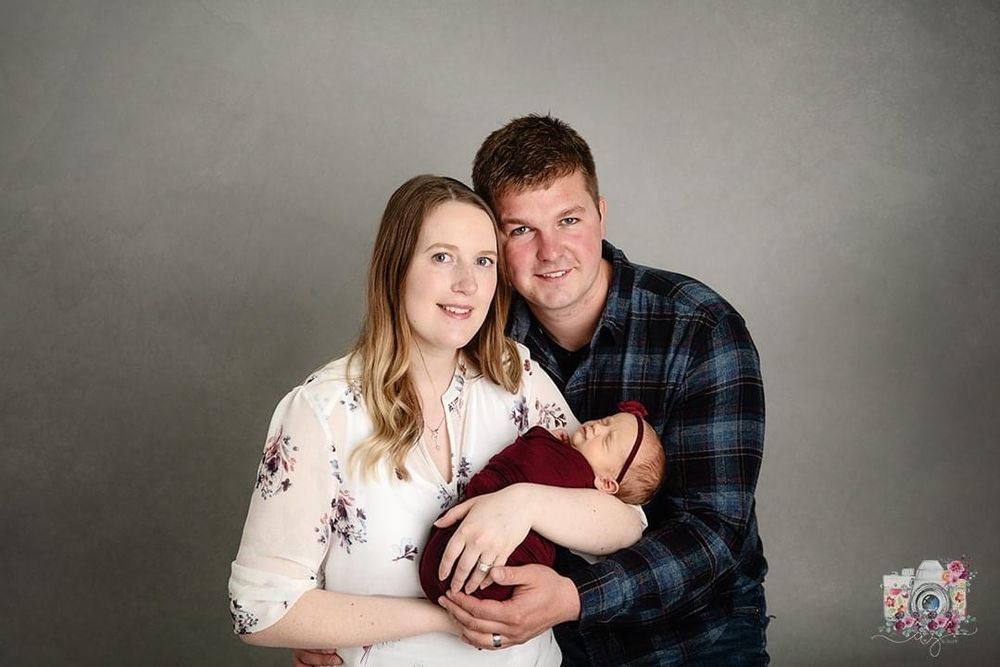 Studio portrait of couple in plaid shirts holding newborn against grey backdrop.