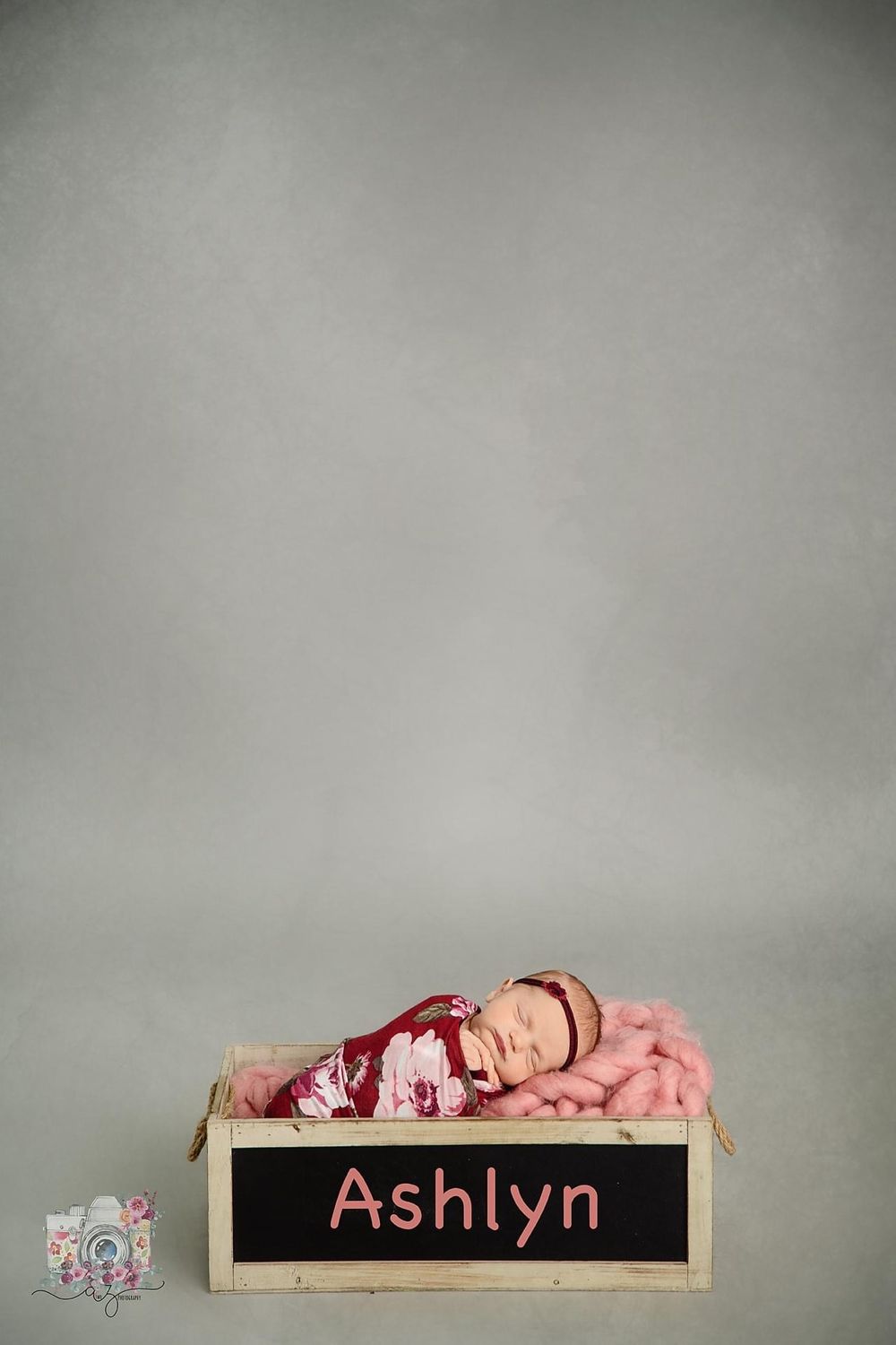 A newborn sleeps peacefully on a pink blanket with their name 'Ashlyn' displayed on a chalkboard below.