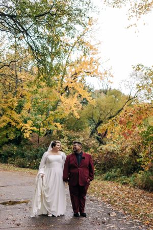 A couple walks along a leaf-covered path surrounded by golden autumn foliage in a park setting.