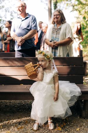 A small child in a white dress sits on a wooden bench holding a basket.
