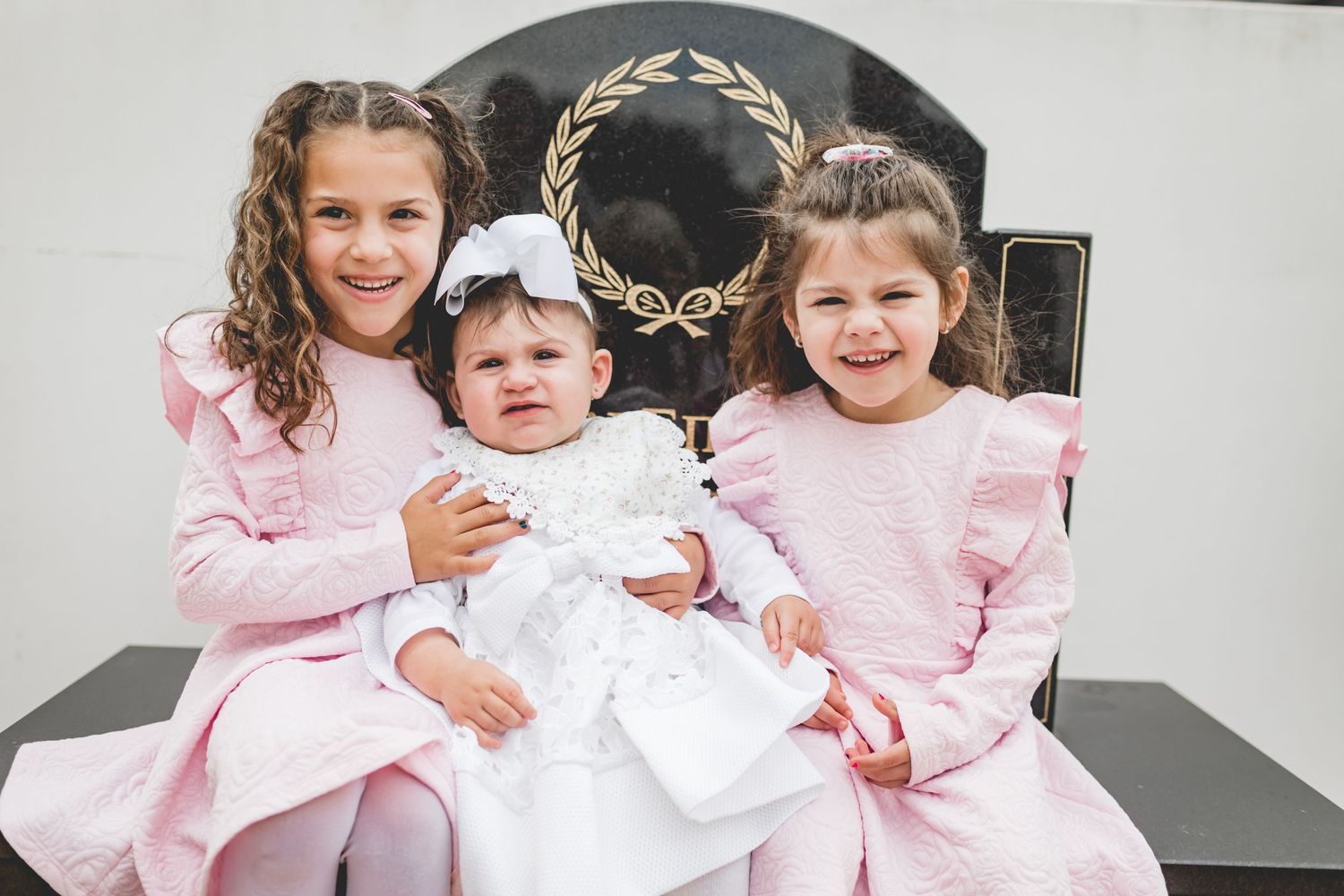Three little girls in pink dresses sitting on a black chair.