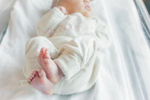 Newborn baby wearing white sleepwear rests peacefully on white bedding.