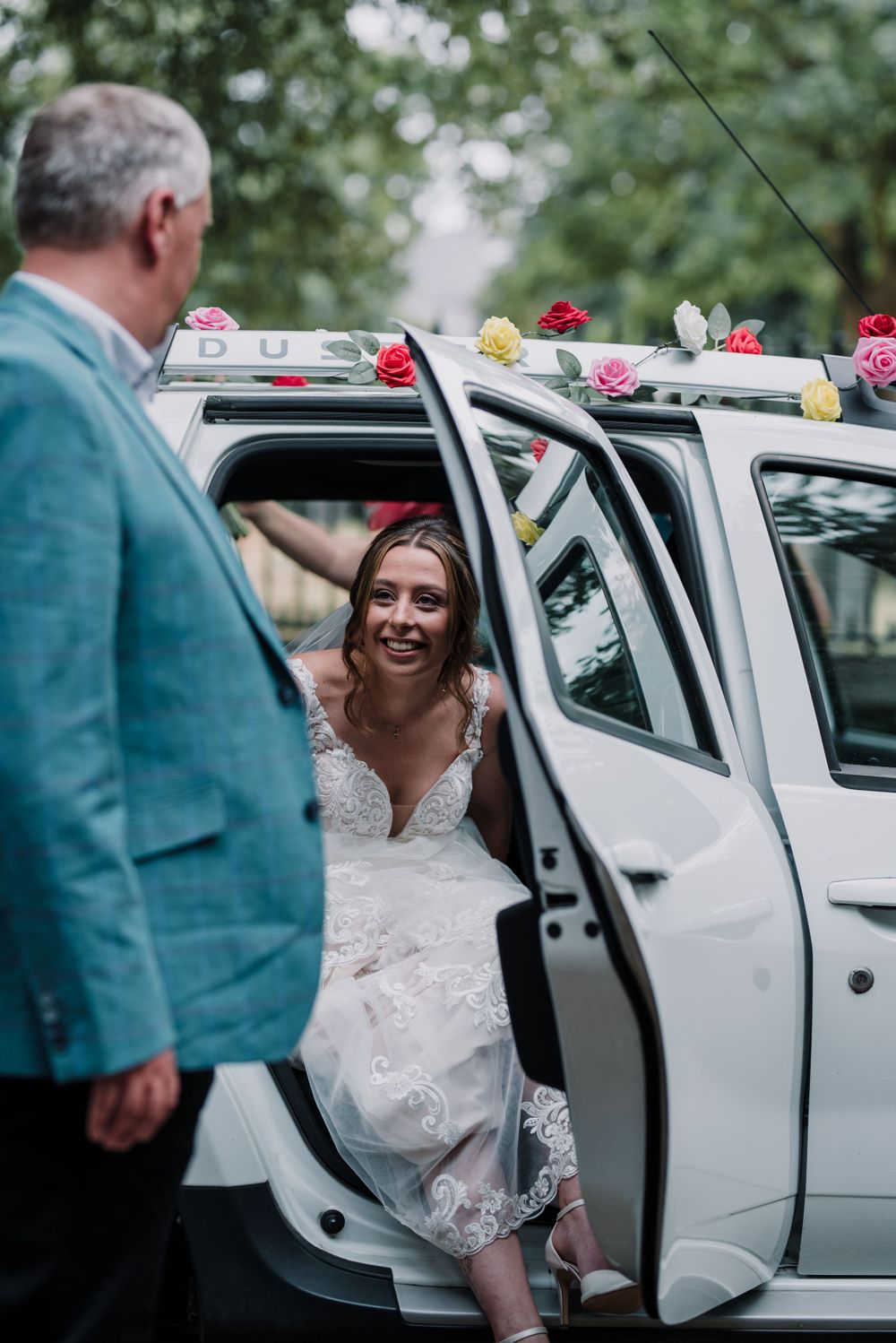 A white wedding car decorated with colorful flowers as someone enters the vehicle.
