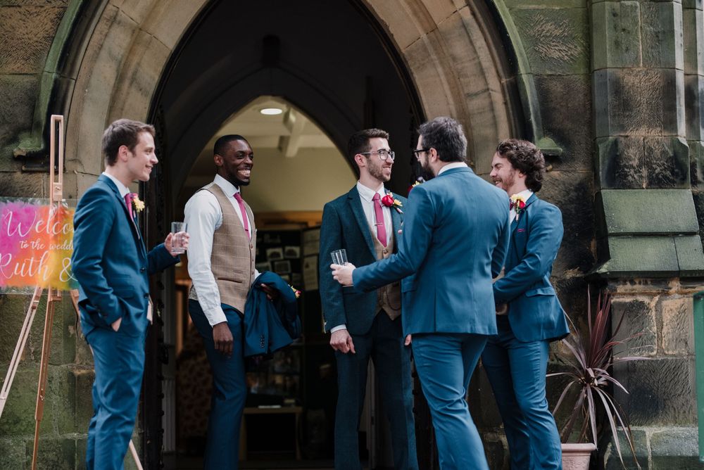 A group of groomsmen in blue suits gather outside a stone archway entrance.