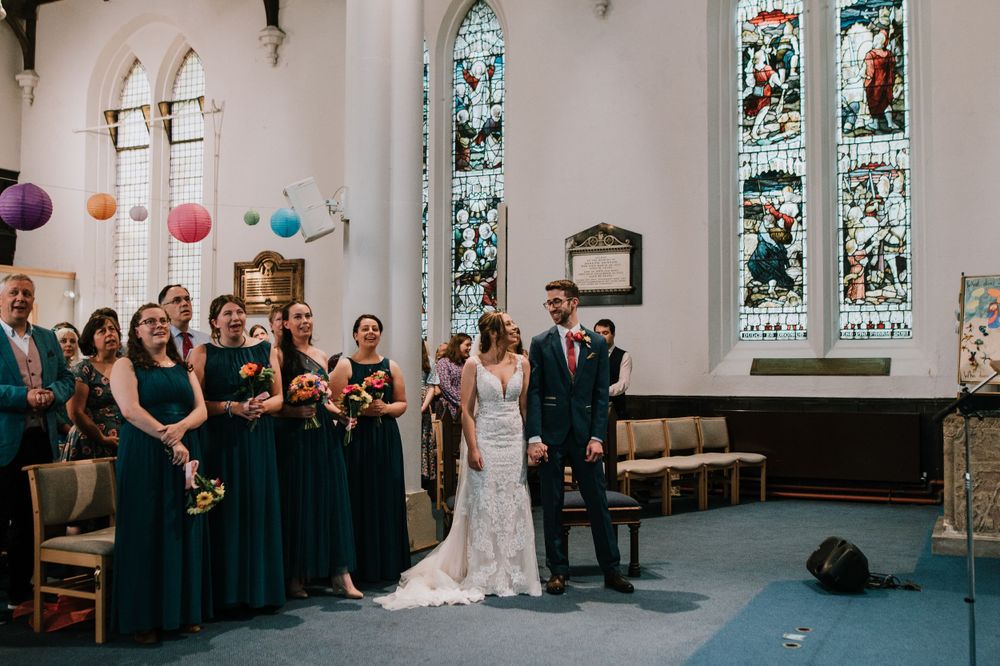 A wedding party stands at the altar in a church with stained glass windows and colorful lanterns.