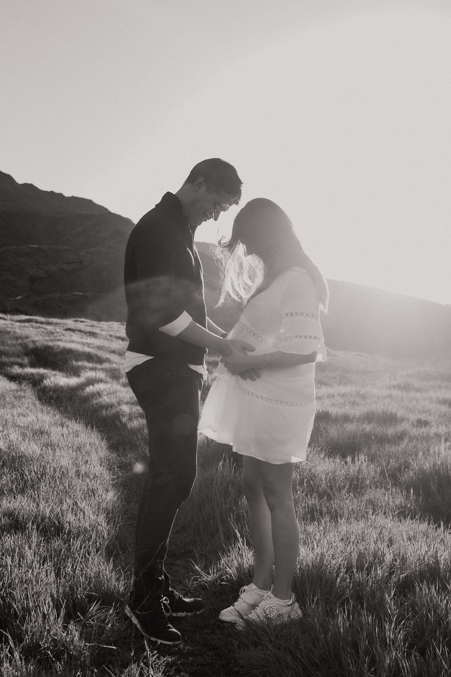 An intimate black and white moment between a couple standing in tall grass with mountains behind them.