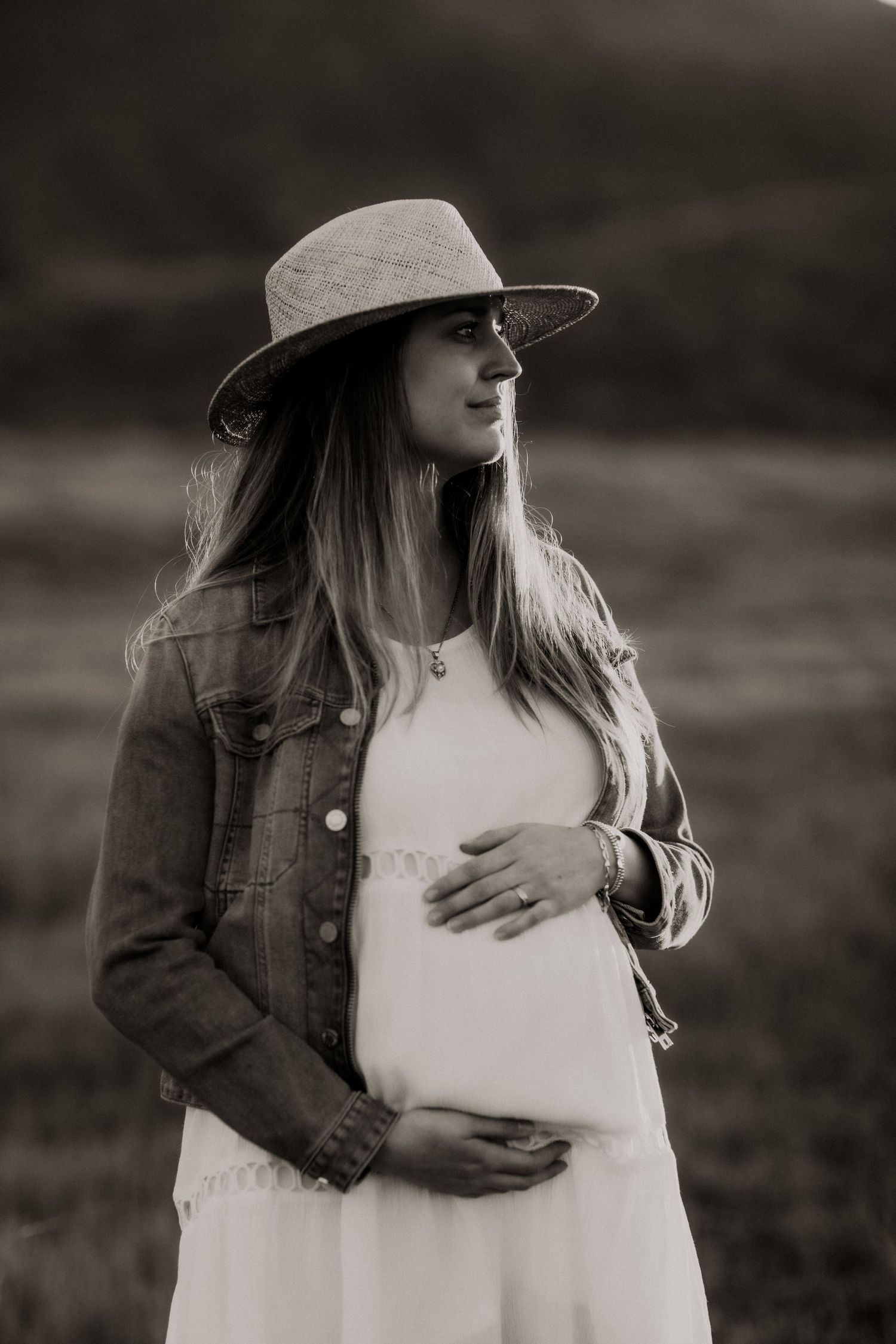 A black and white portrait of an expectant mother wearing a wide-brimmed hat and denim jacket.
