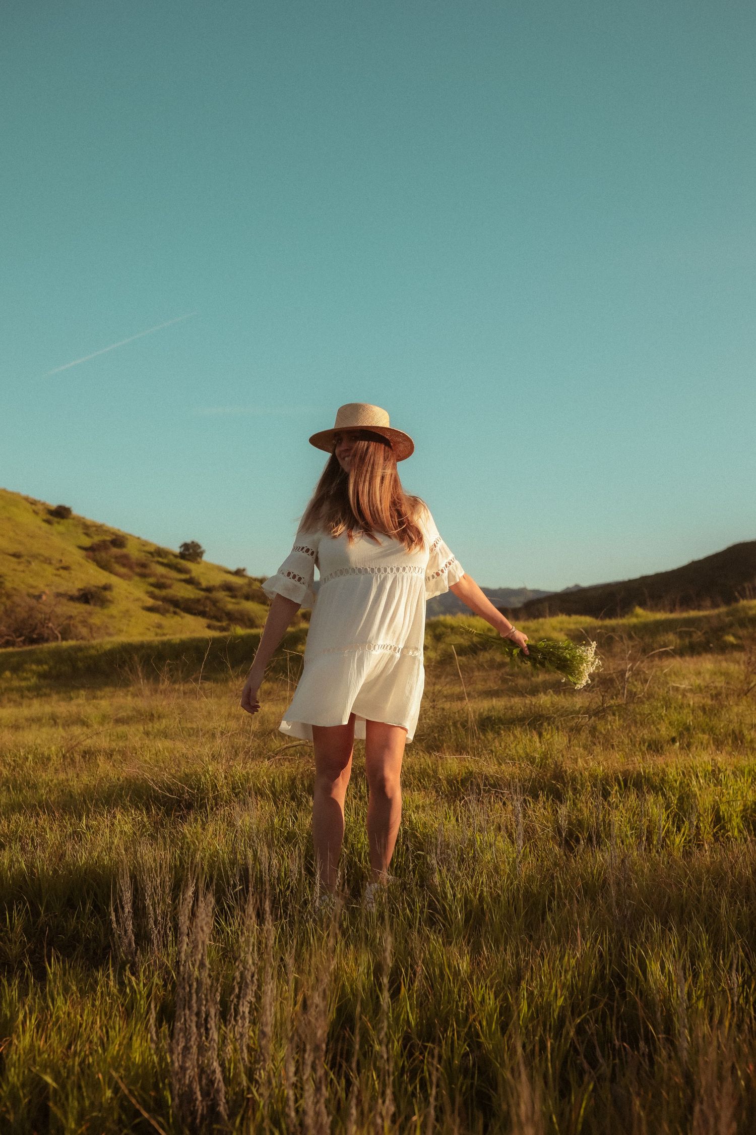 A figure in a flowing white dress twirls through a sunlit meadow against rolling hills.