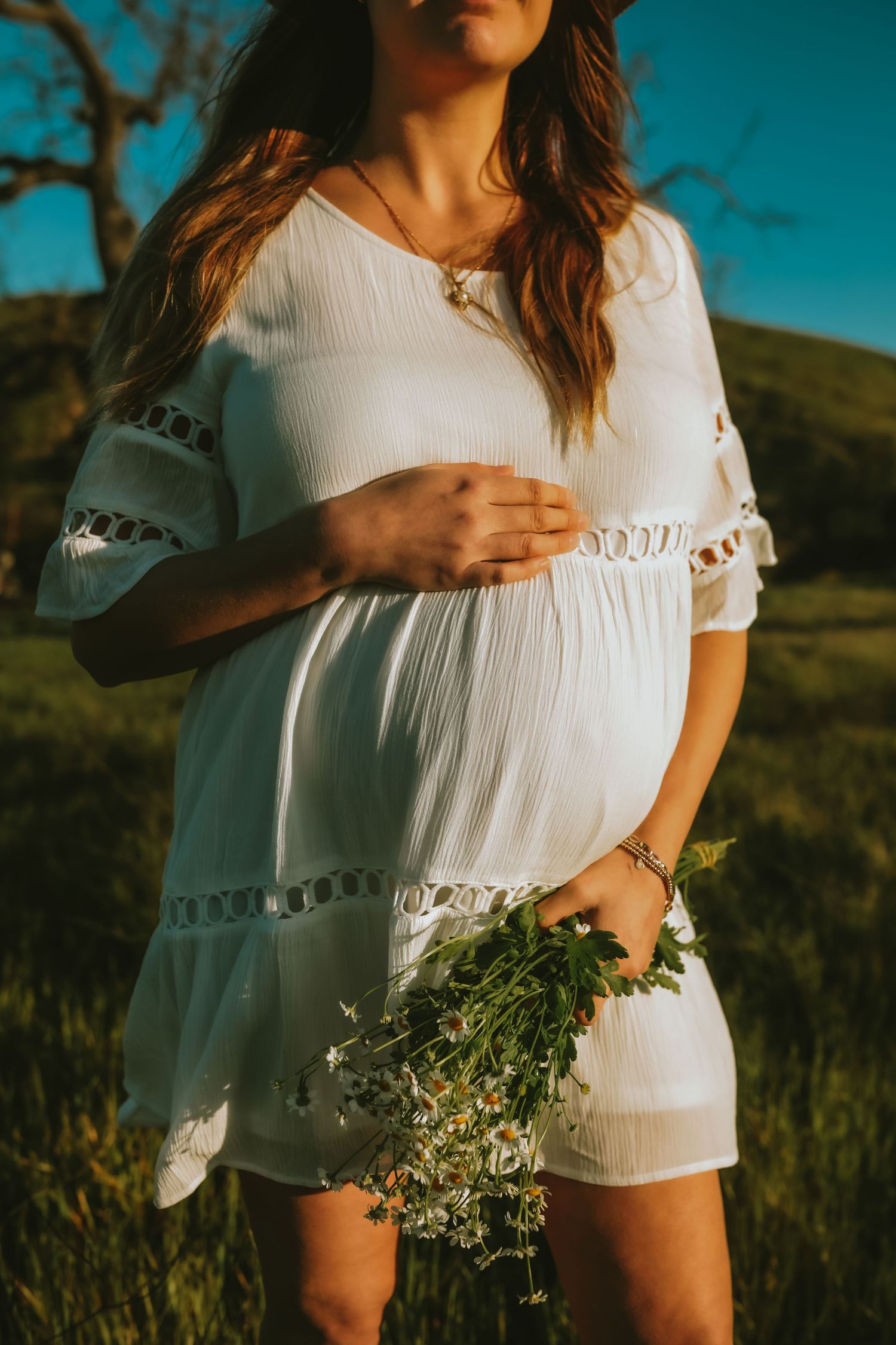 A pregnant woman in a white lace dress holds wildflowers during golden hour in a scenic field.