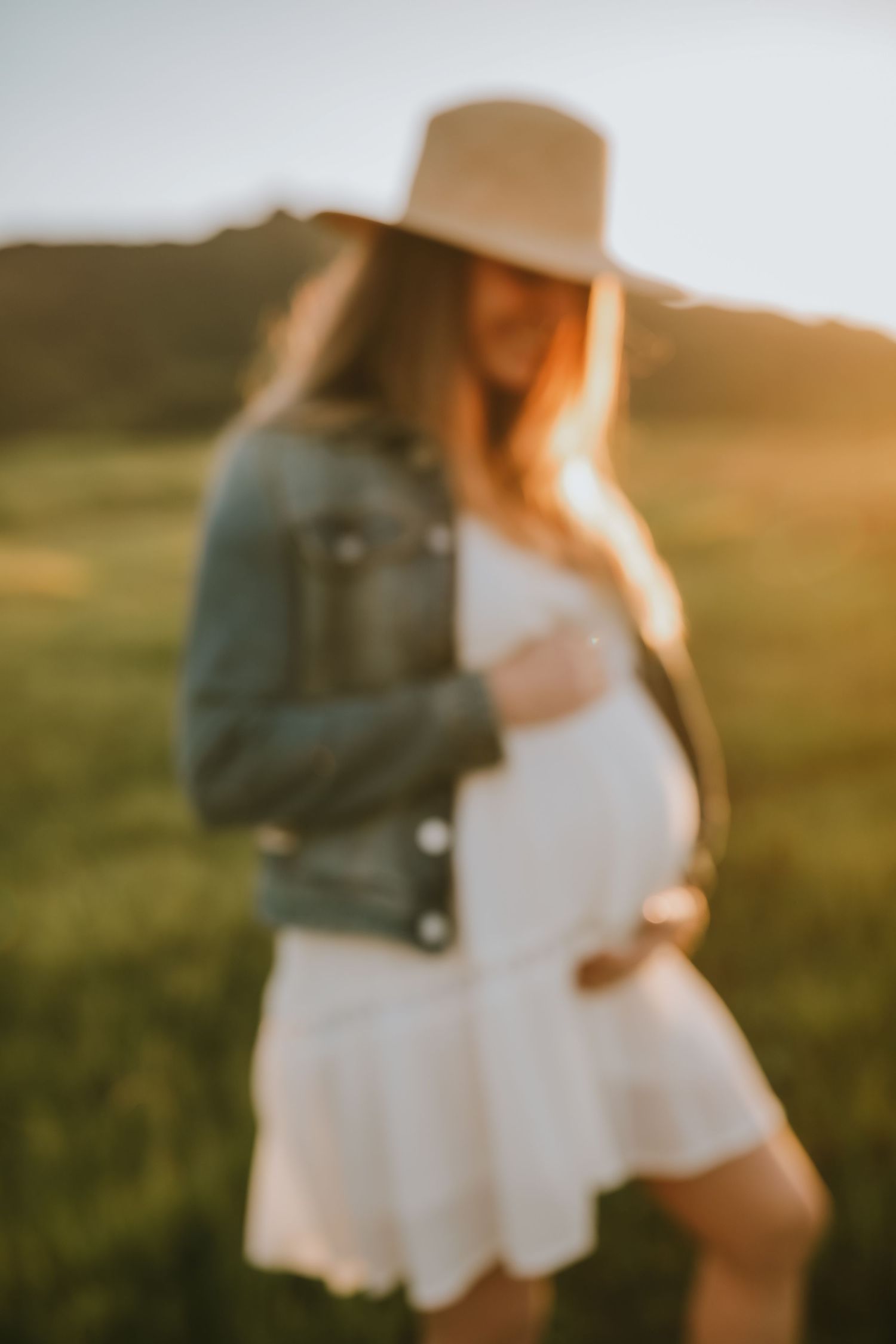A dreamy sun-lit maternity photo showing a woman in a white dress and denim jacket standing in a grassy field.