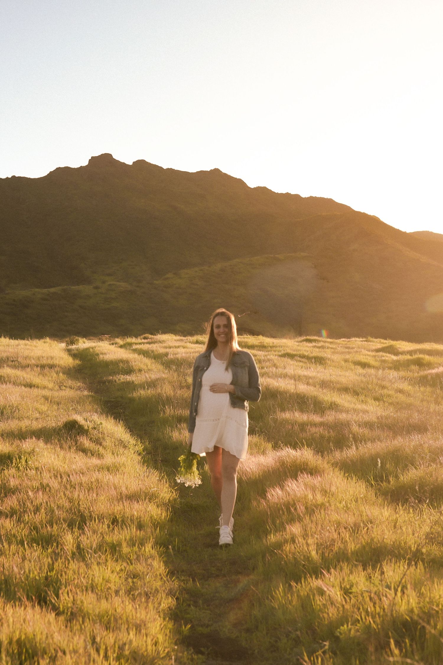 Figure in white dress and denim jacket walks through sun-drenched mountain meadow.