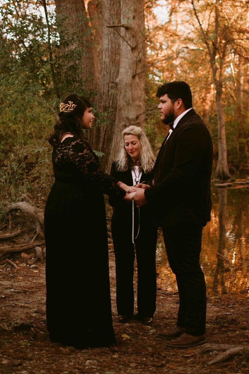 Three people stand together during an intimate forest wedding ceremony with reflective water nearby.
