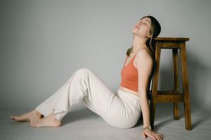 Relaxed studio pose in coral tank top and white pants next to wooden stool.