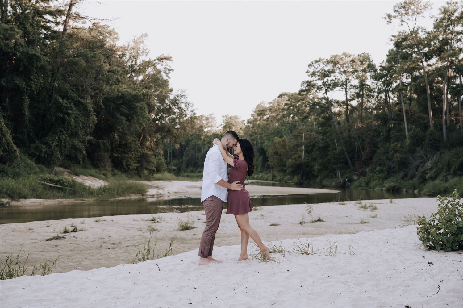 Couple sharing an intimate moment on a sandy riverbank surrounded by forest.
