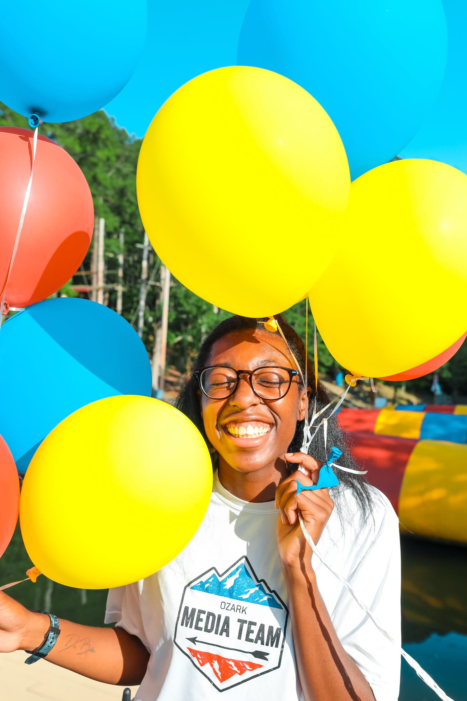 Kassidy/KLo holding yellow. blue, and red balloons and smiling in front of a waterfront.