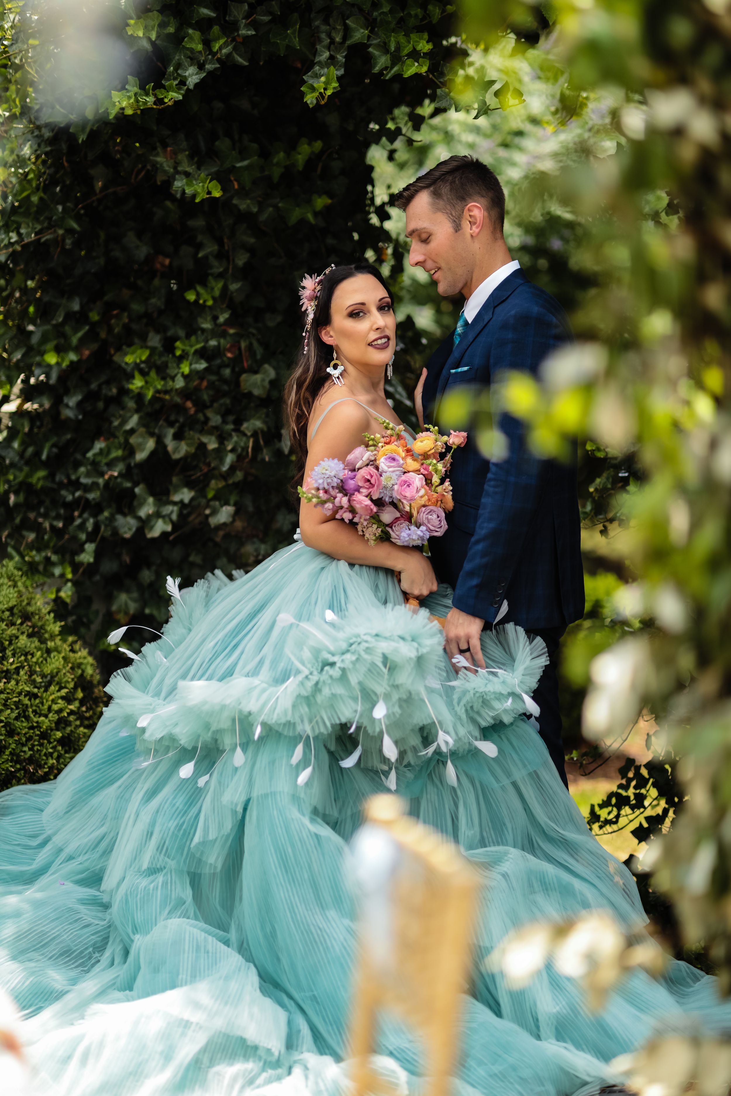 Couple in formal attire poses beneath greenery with bride wearing flowing mint green dress and holding colorful bouquet.