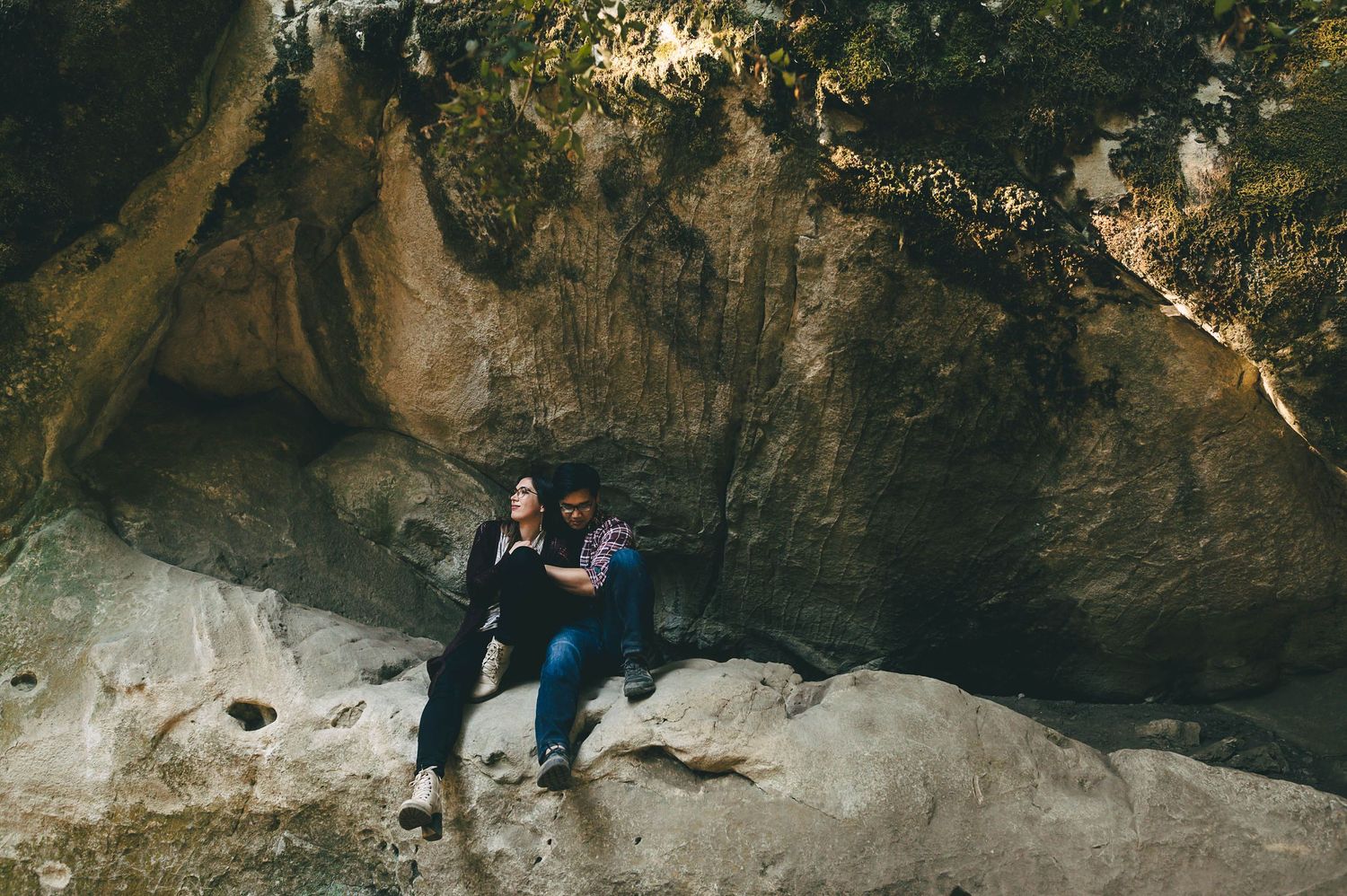 Person sitting on rocky outcrop beneath large cave entrance in nature.