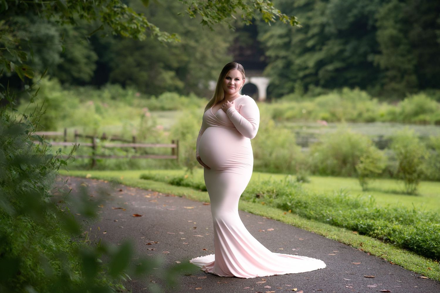 Ethereal maternity portrait in a flowing pink gown against a lush garden path backdrop.
