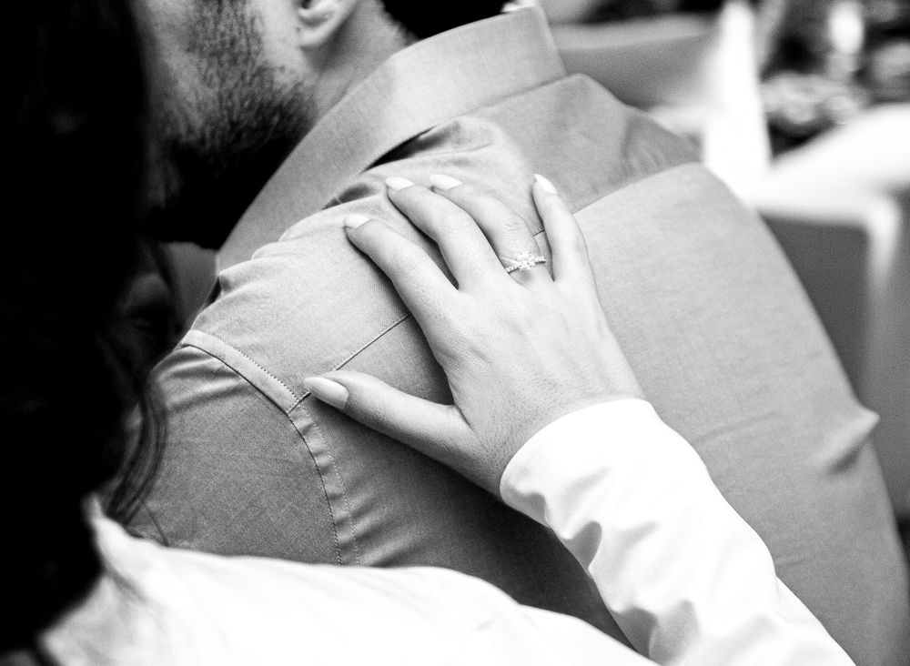 Black and white closeup of hands wearing an engagement ring embracing someone in formal wear.