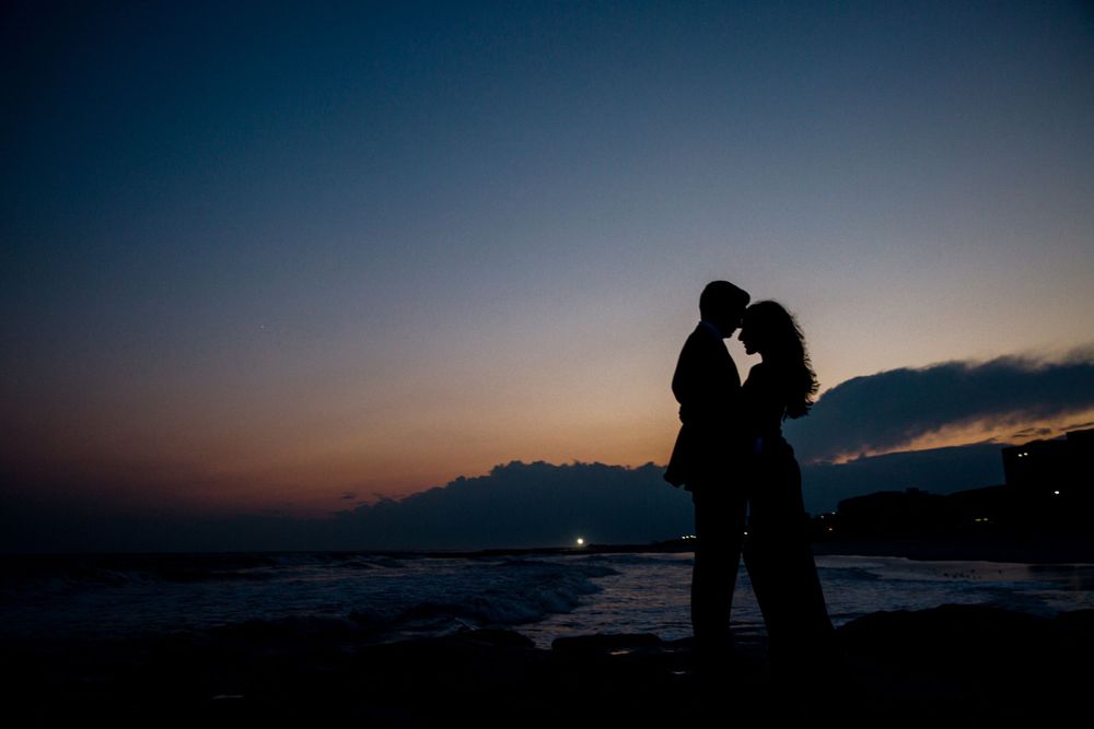 Silhouetted couple embracing against dramatic sunset sky by ocean.