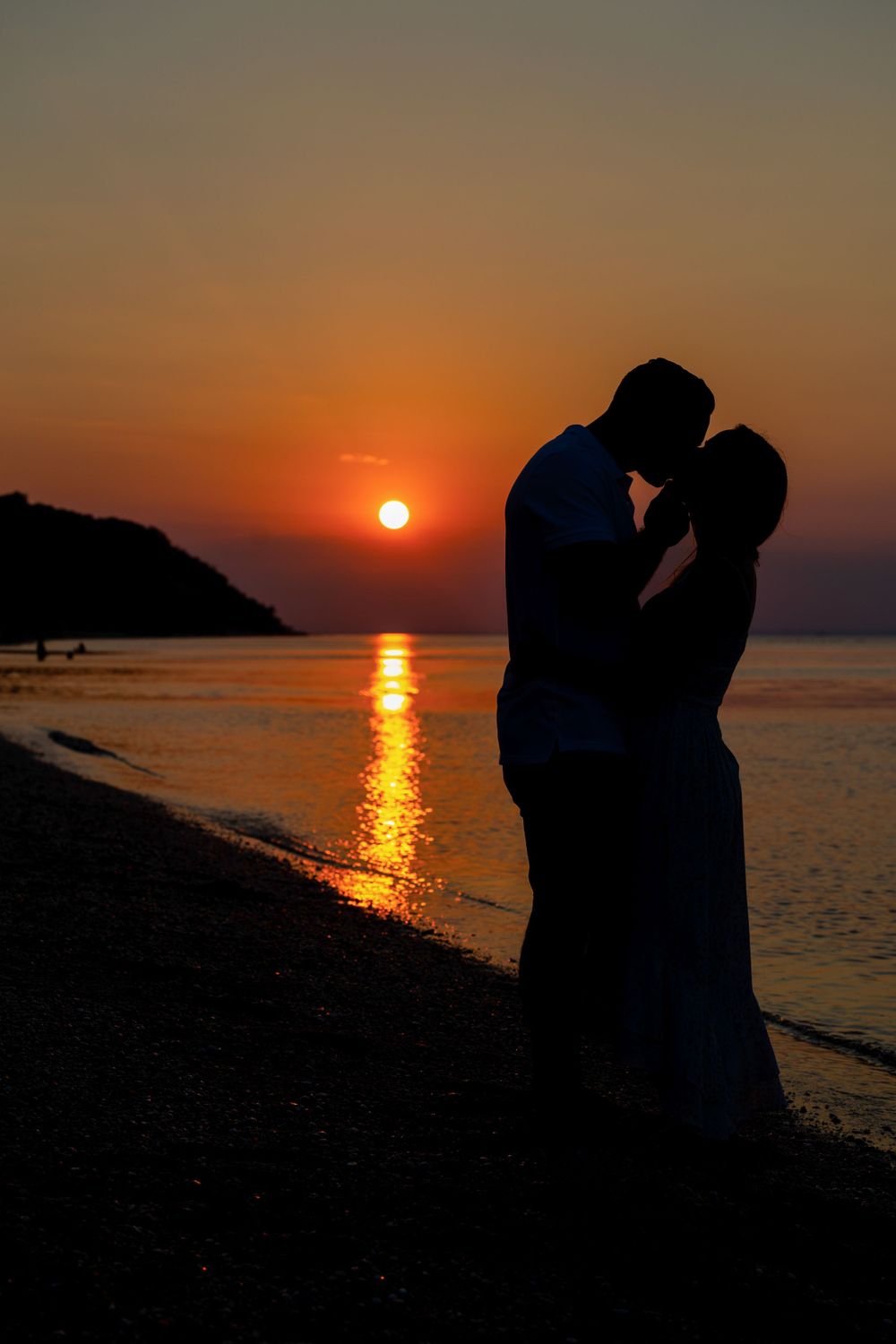 A silhouetted couple sharing a romantic moment against a vibrant orange sunset over the ocean.