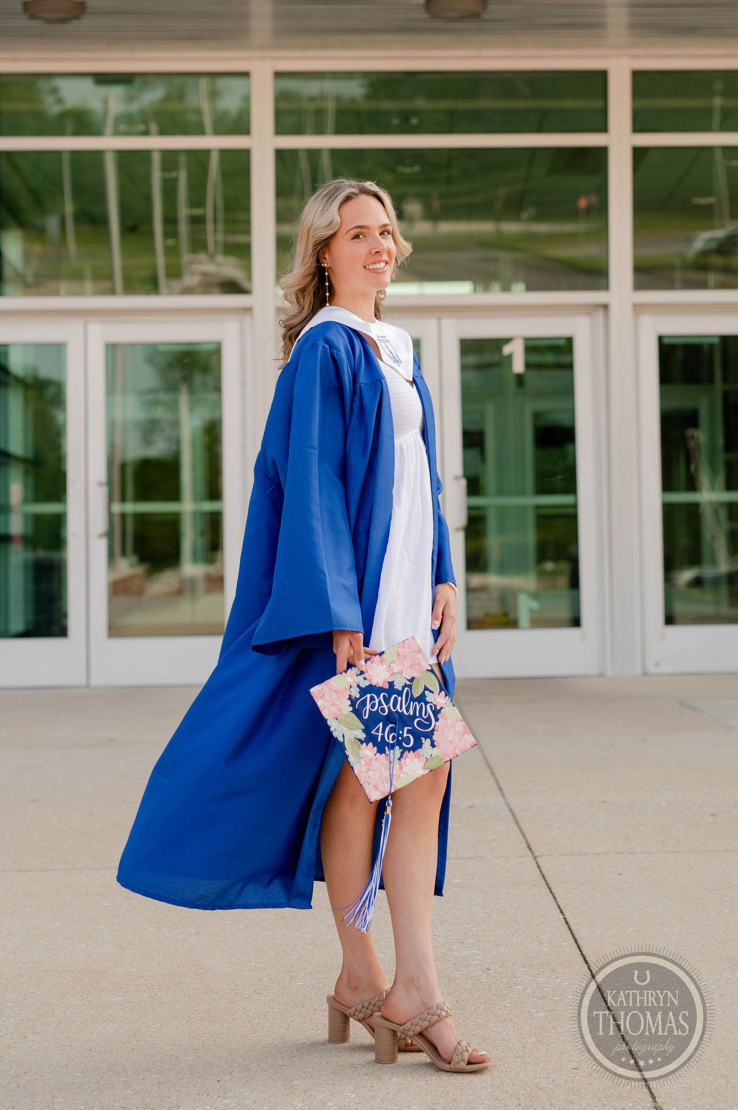 Graduate wearing white dress and blue gown holds decorated graduation cap outside school building.