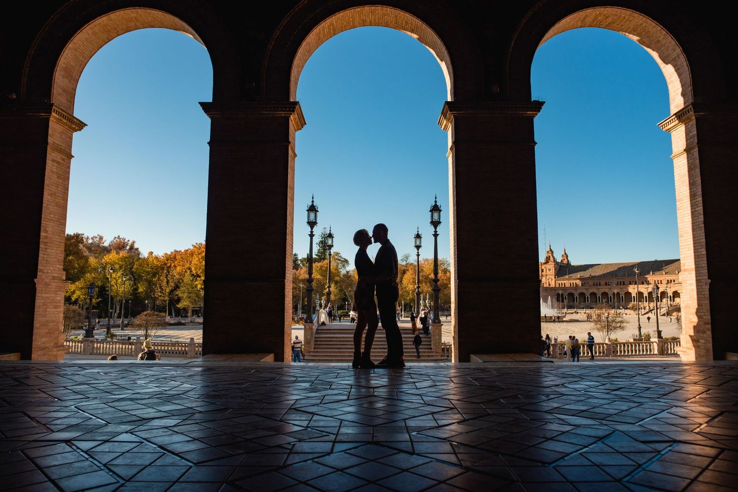 Silhouetted figures stand in arched colonnade overlooking Plaza de España.