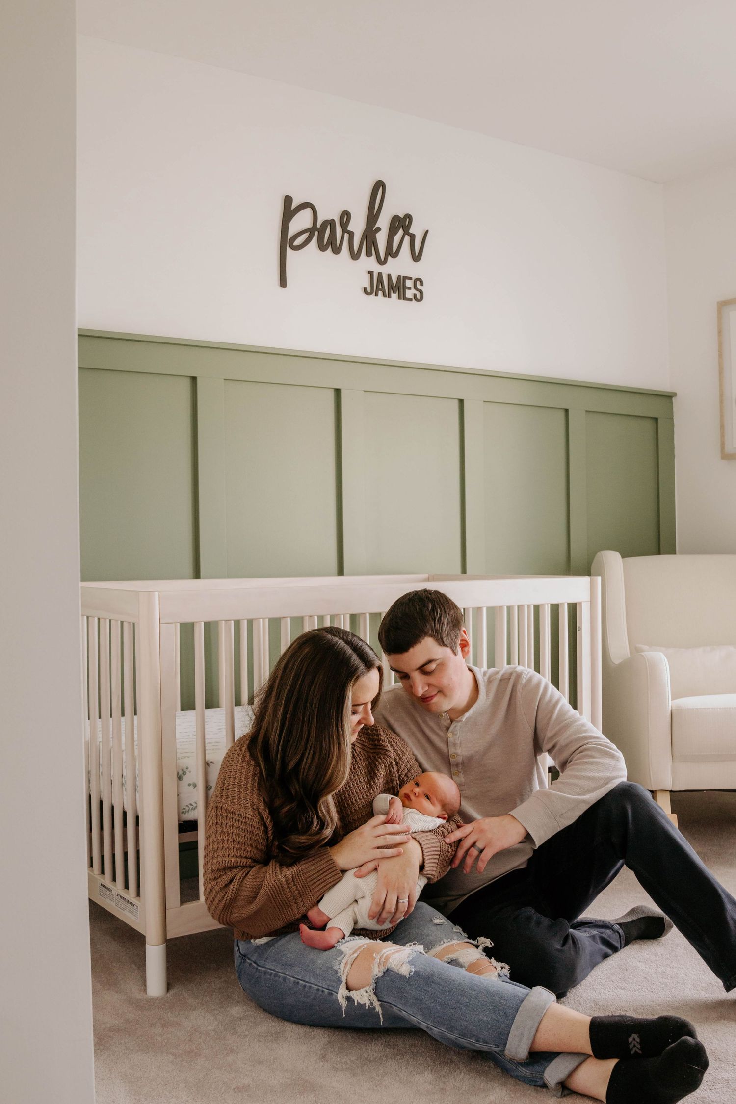 New parents sitting on nursery floor holding newborn beneath Parker James name sign by crib.