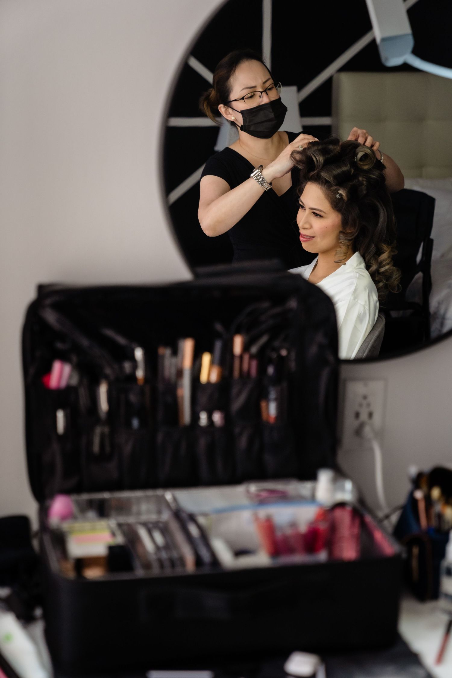 Hair stylist in black mask styling brunette woman's hair in salon with makeup kit in foreground.