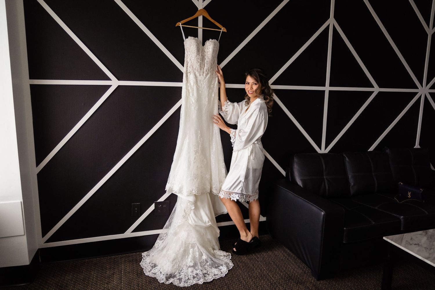 Bride in white robe admiring her lace wedding gown hanging on a hanger in a modern hotel suite.