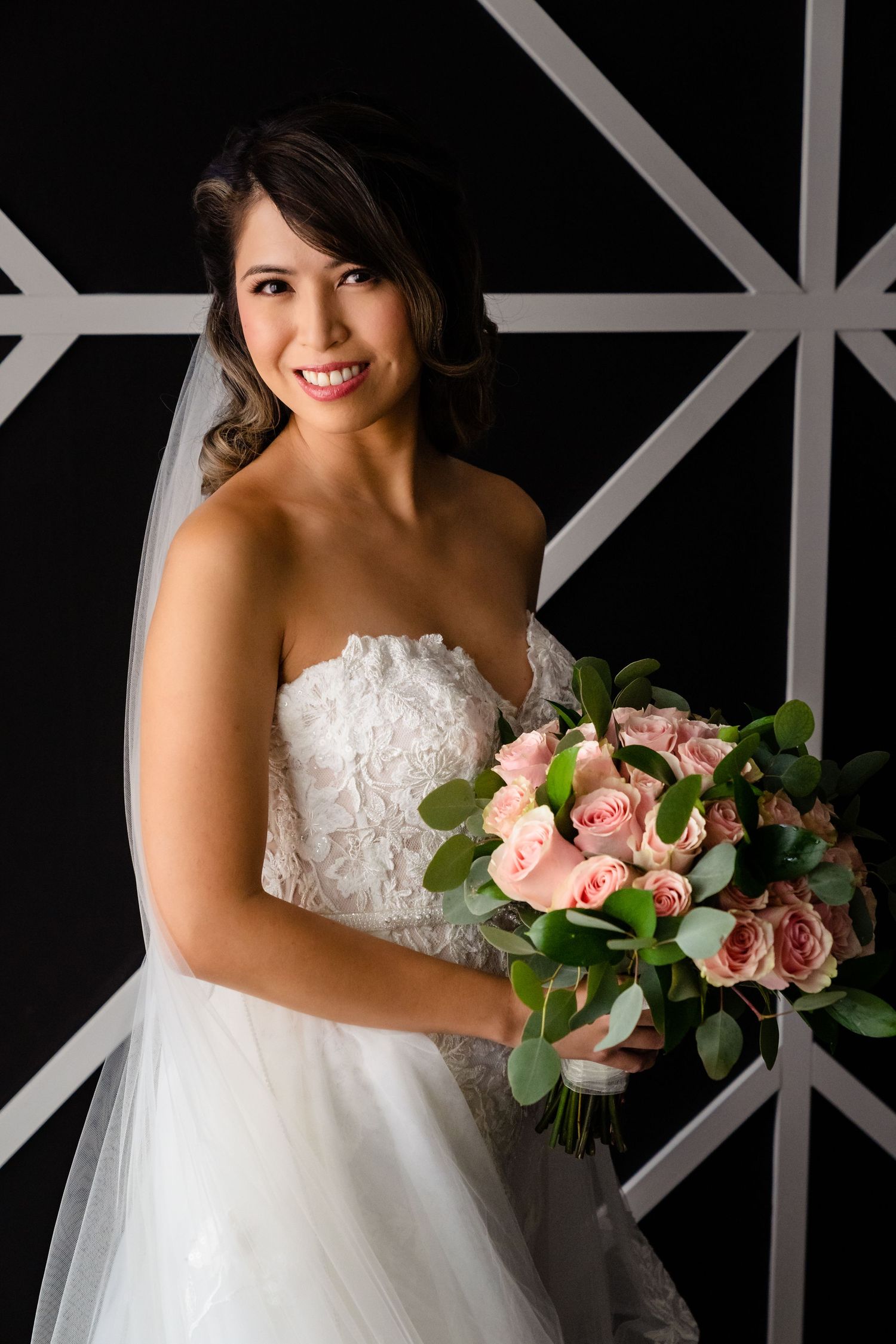 Elegant bride in strapless lace gown holding pink rose bouquet against geometric black and white wall.