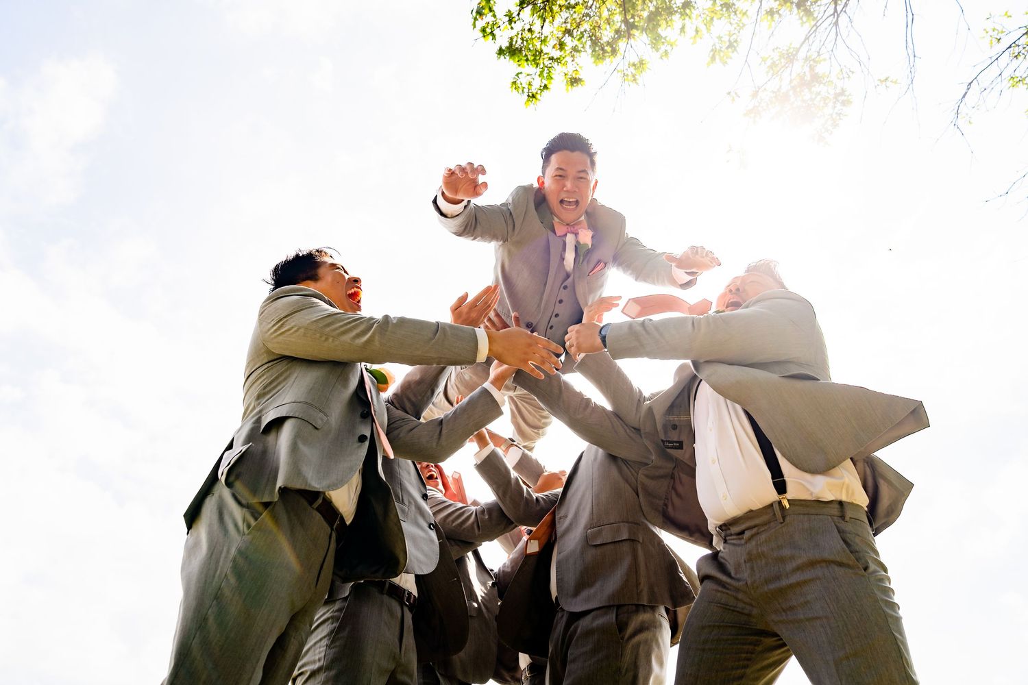 Groomsmen lifting groom in celebratory pose against bright sky.