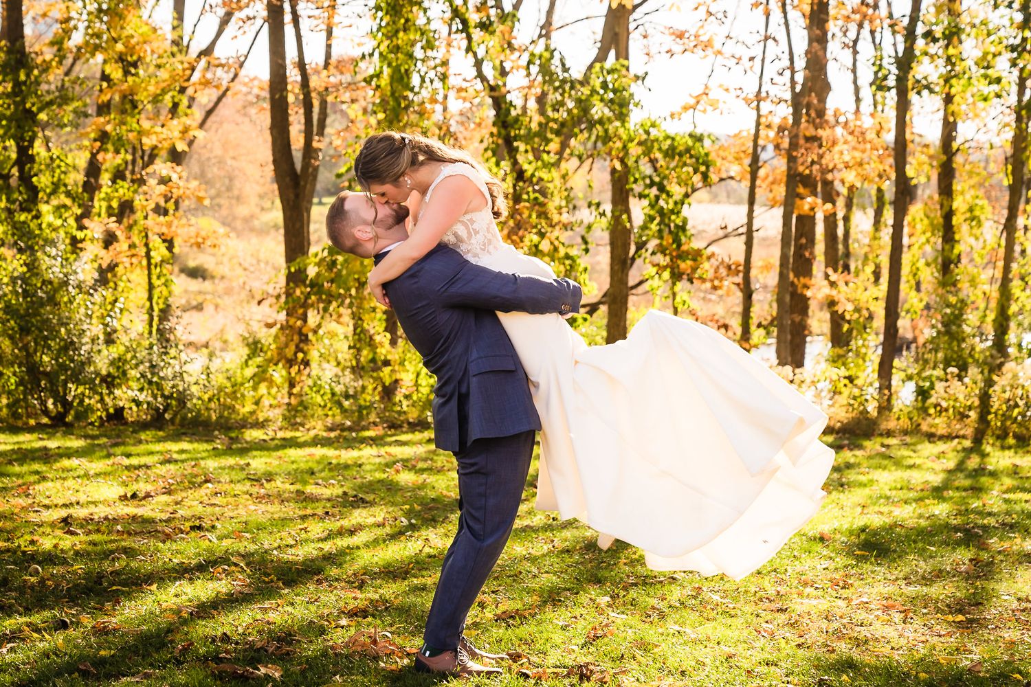 Groom lifts bride in white dress during outdoor fall wedding photo among golden autumn trees.