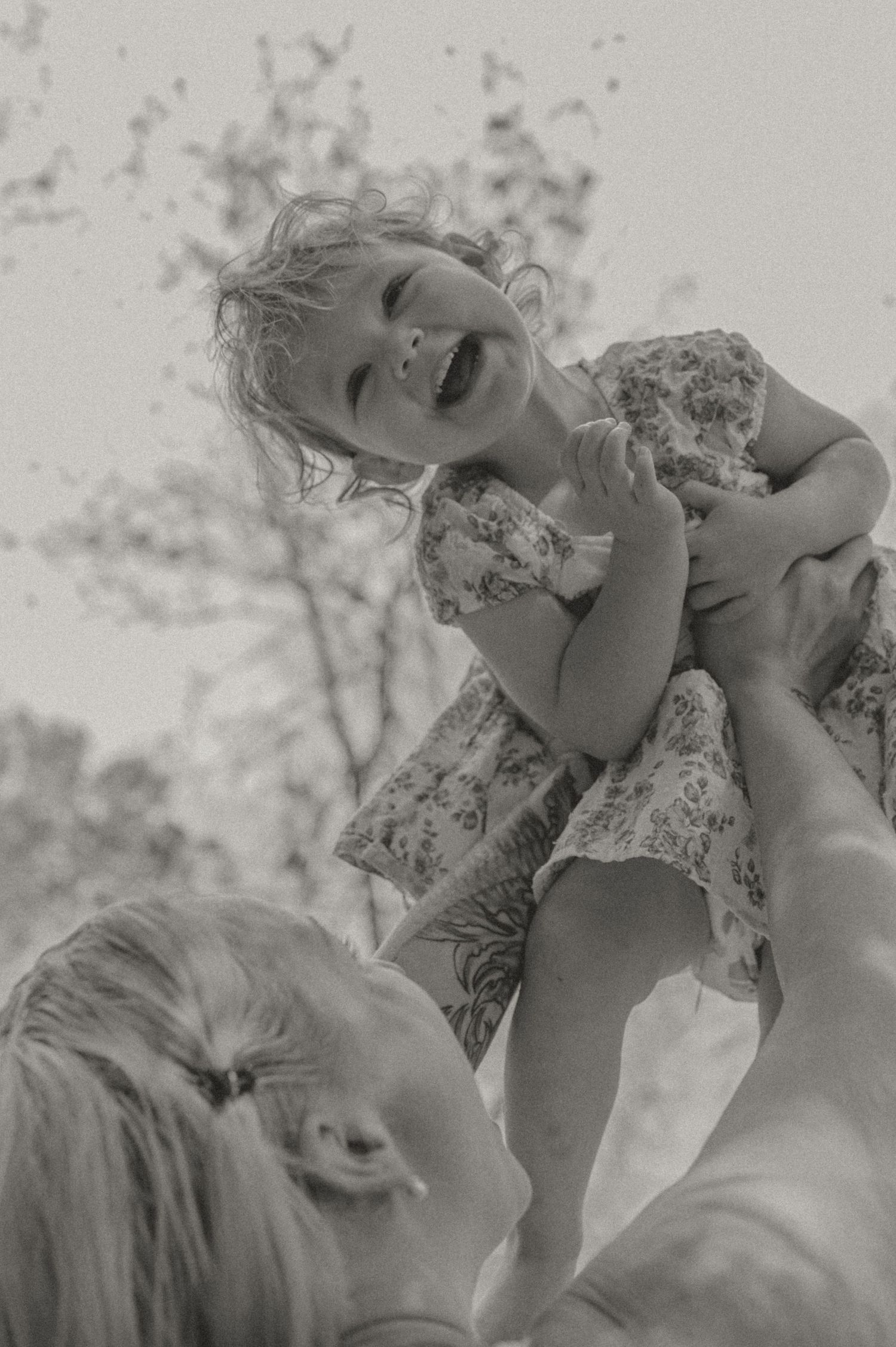 Black and white photograph of joyful parent lifting a small child wearing a floral dress against a blurred outdoor background.