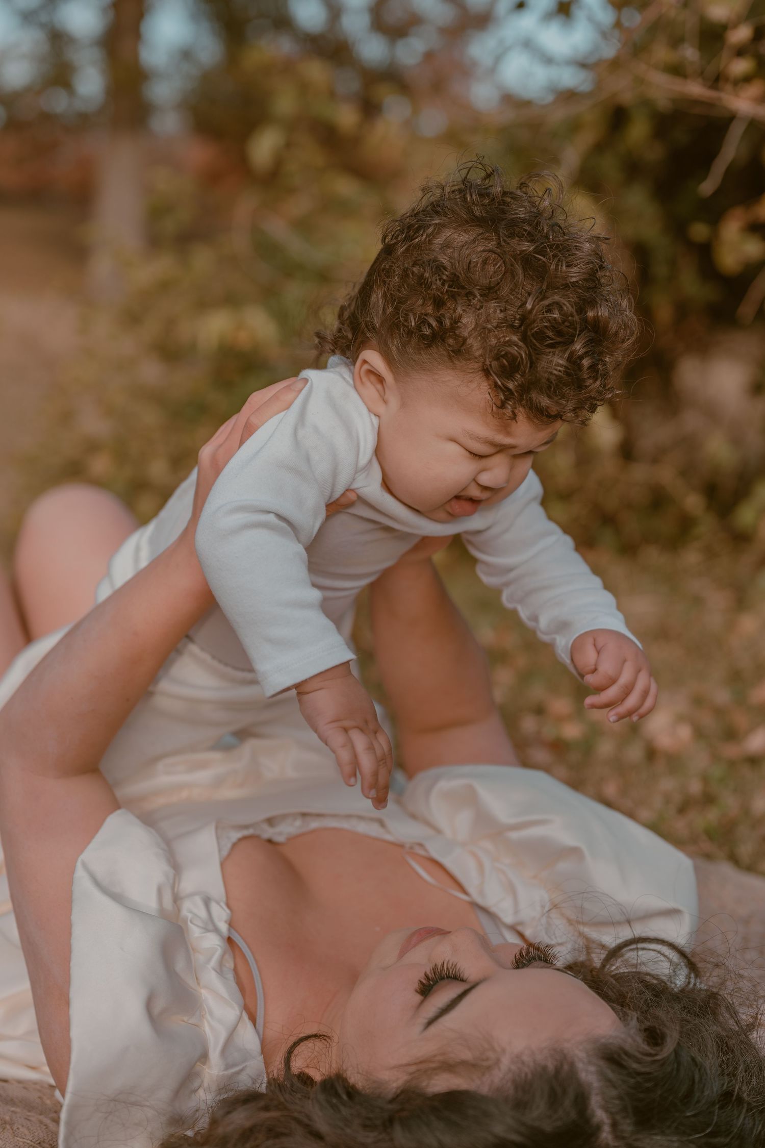 A mother lying on the ground outdoors, playfully holding her curly-haired baby in a white onesie amid autumn foliage.