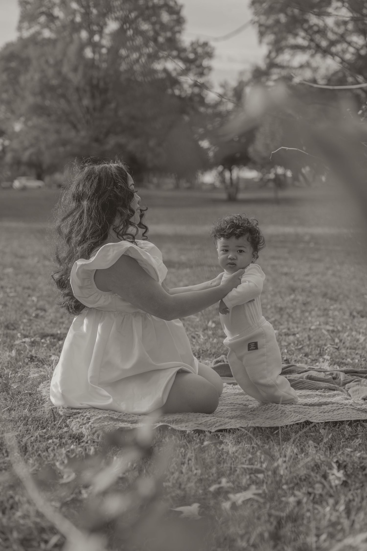 Black and white photo of a mother in white dress playing with her toddler on a blanket in a lush green park.
