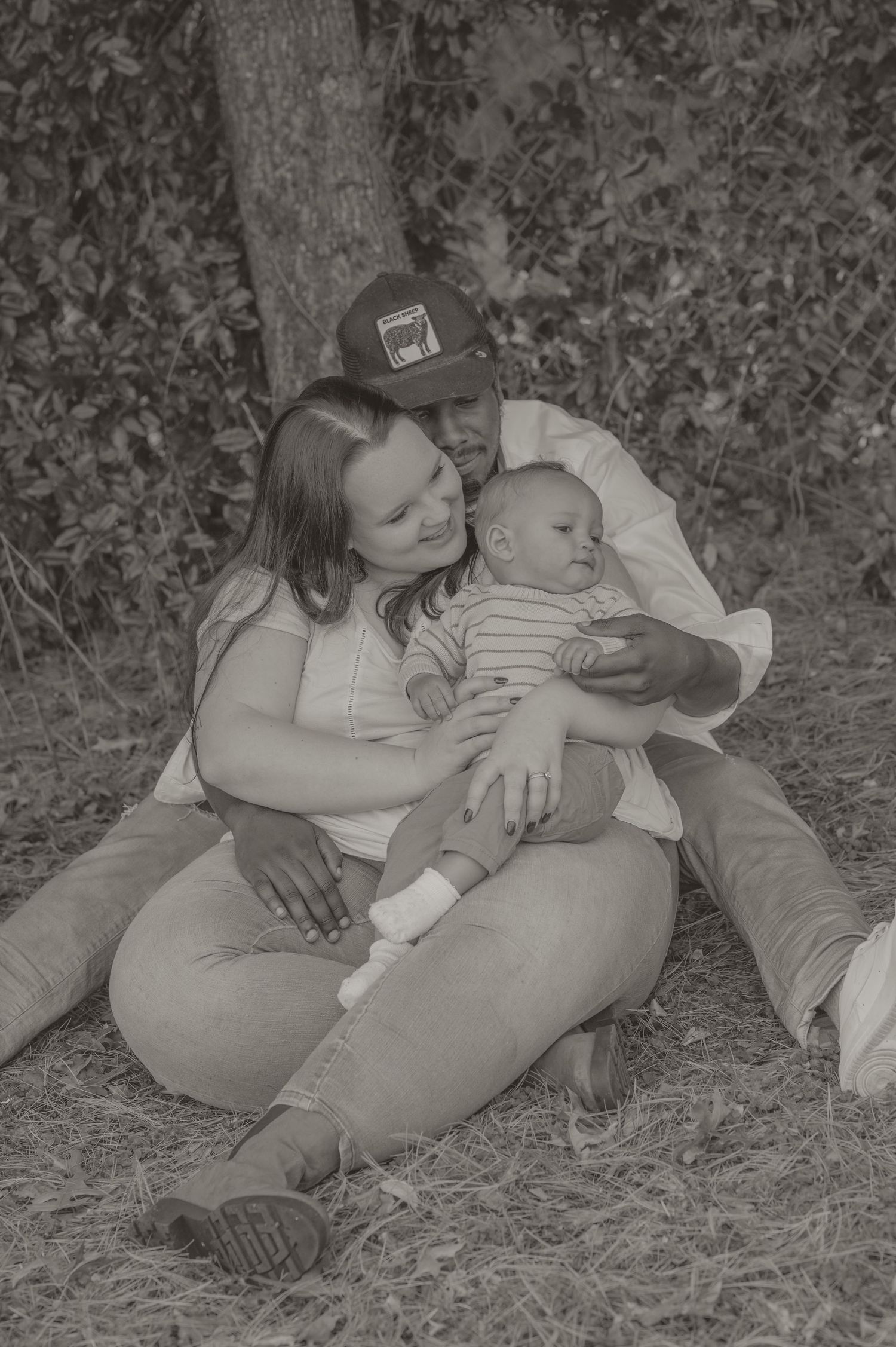 Black and white photo of a couple sitting outdoors cradling their newborn baby near trees.