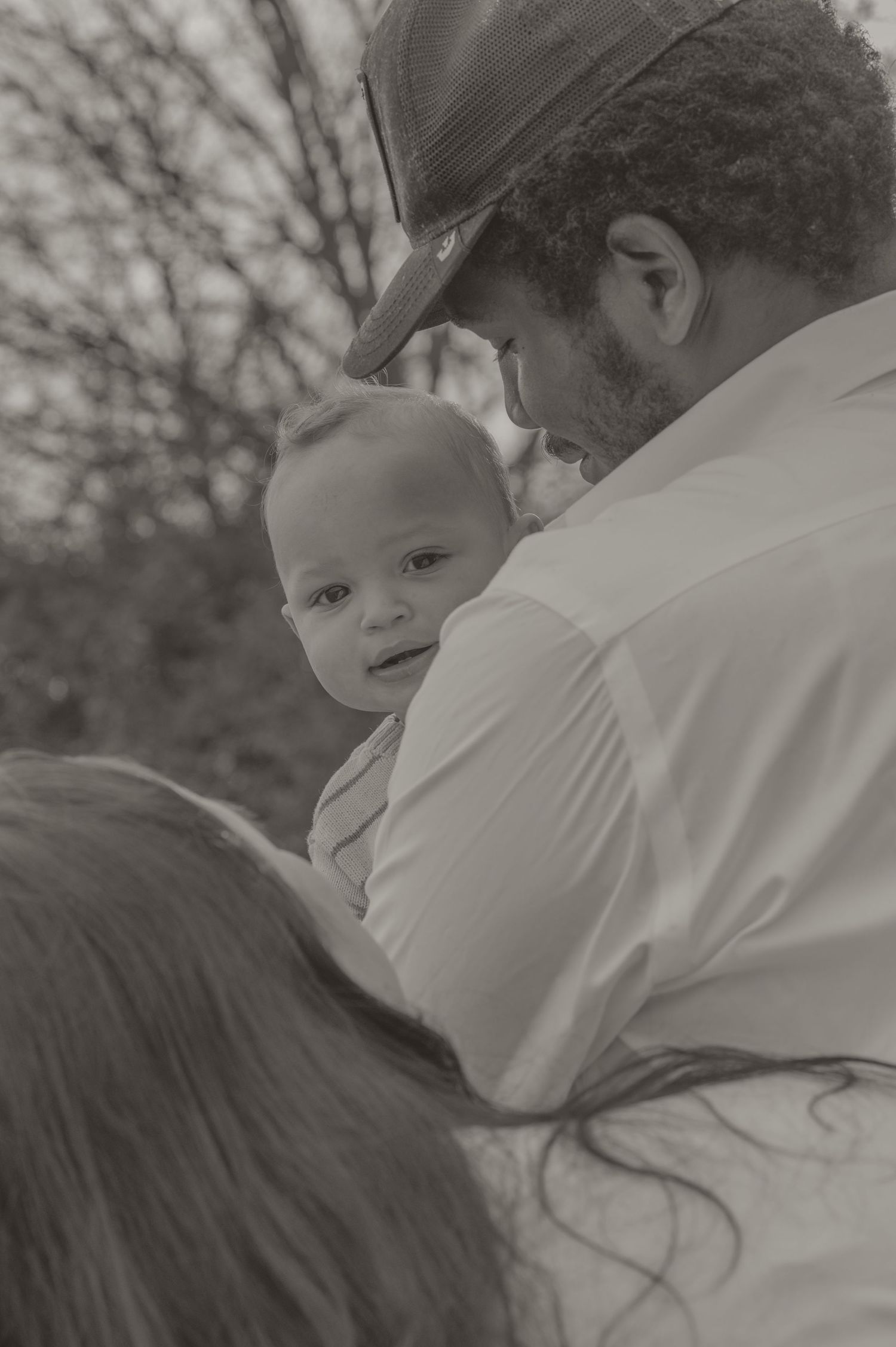 Black and white photo of a man in a cap tenderly holding a baby outdoors with bare trees in the background.