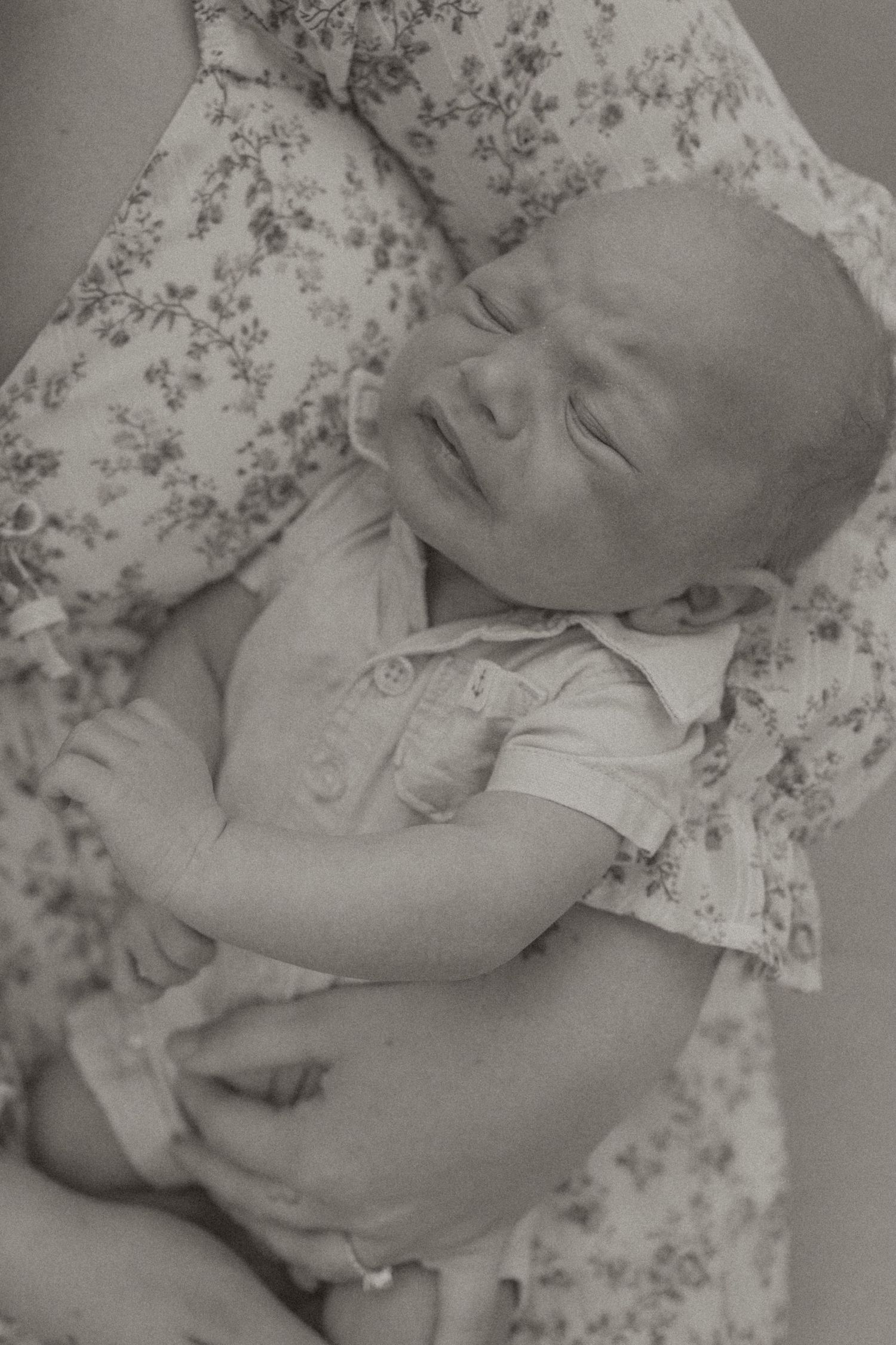 Black and white photo of a sleeping newborn baby cradled in loving arms, wearing a onesie with floral fabric background.