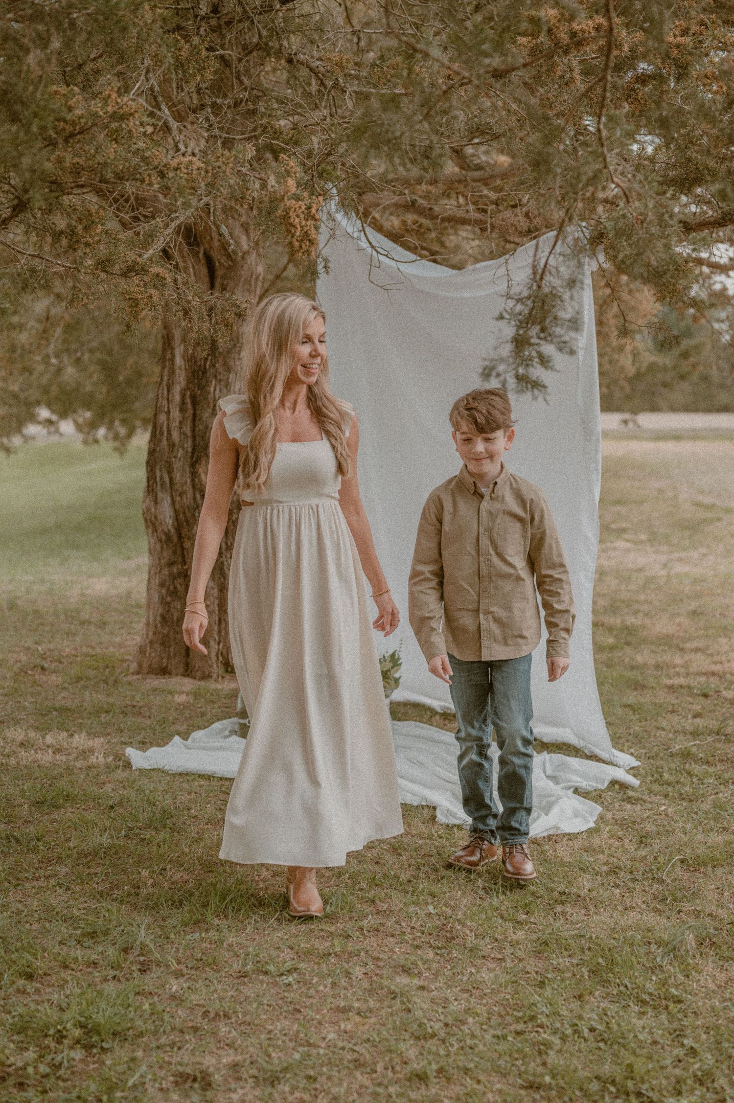 Two figures in casual attire pose together in front of a white sheet backdrop hanging from a tree in a park setting.