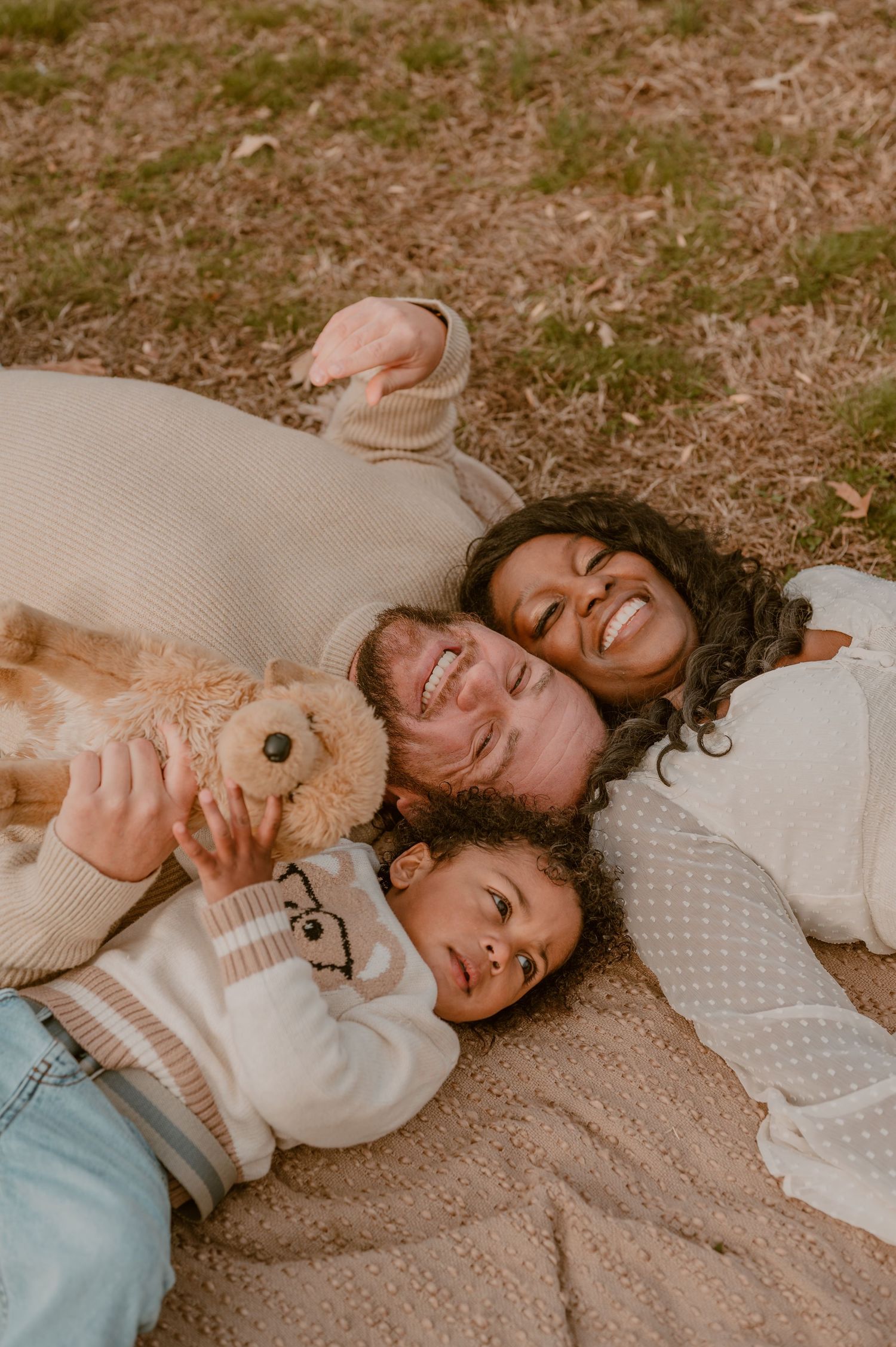 Three people relaxing together on a blanket outdoors on the grass in warm sweaters with a teddy bear.