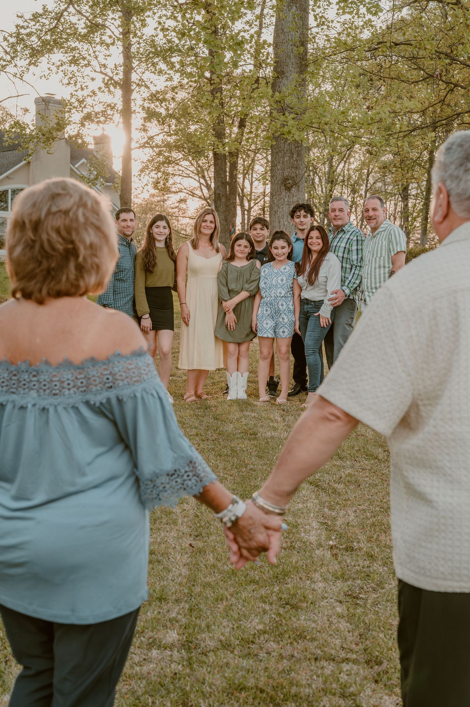 A group gathers on a grassy lawn at sunset while a couple holds hands in the foreground among trees.