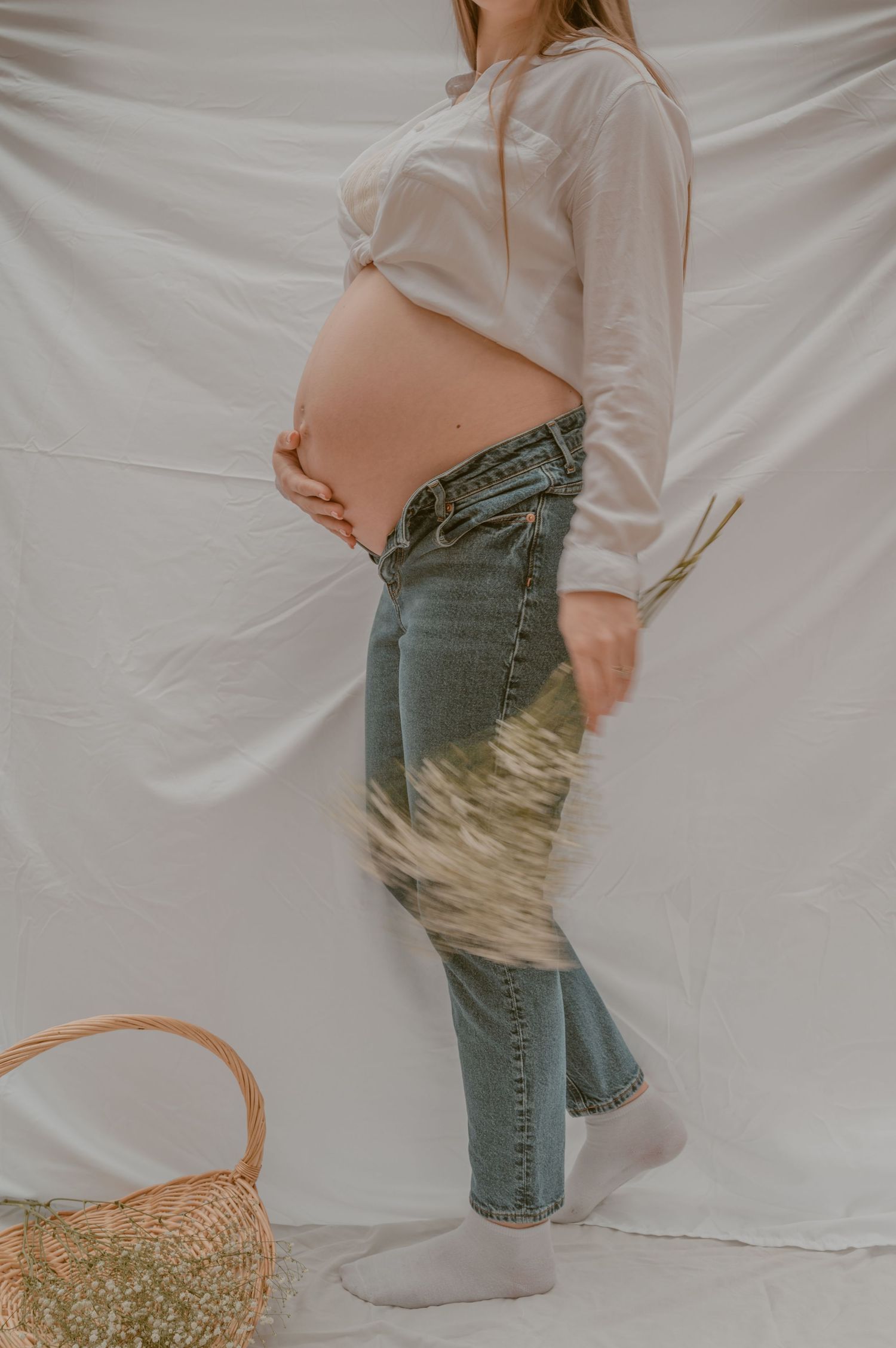 Side view of pregnant belly in casual outfit with white button down shirt and jeans against white backdrop with straw basket.