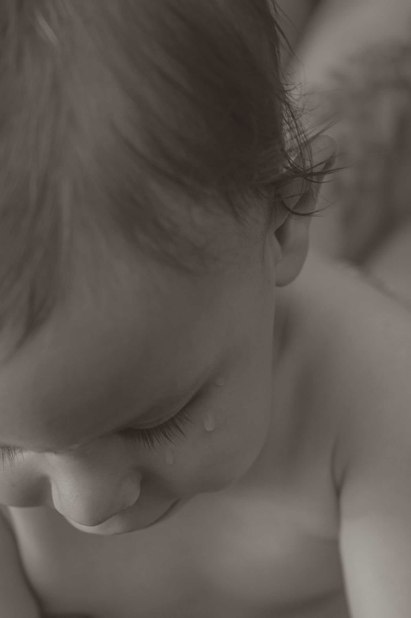 Black and white close-up portrait of a crying baby with a tear on cheek, looking downward.