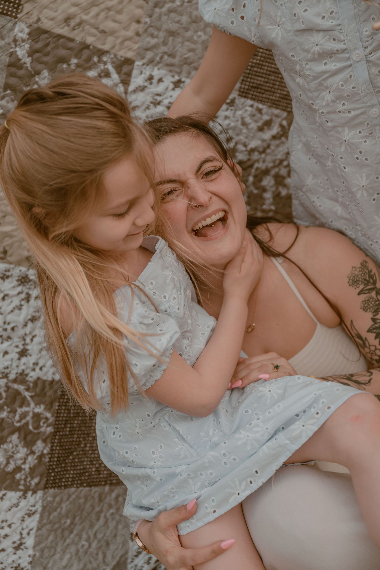 A laughing mother and her young blonde daughter share a joyful moment together on a patchwork quilt.