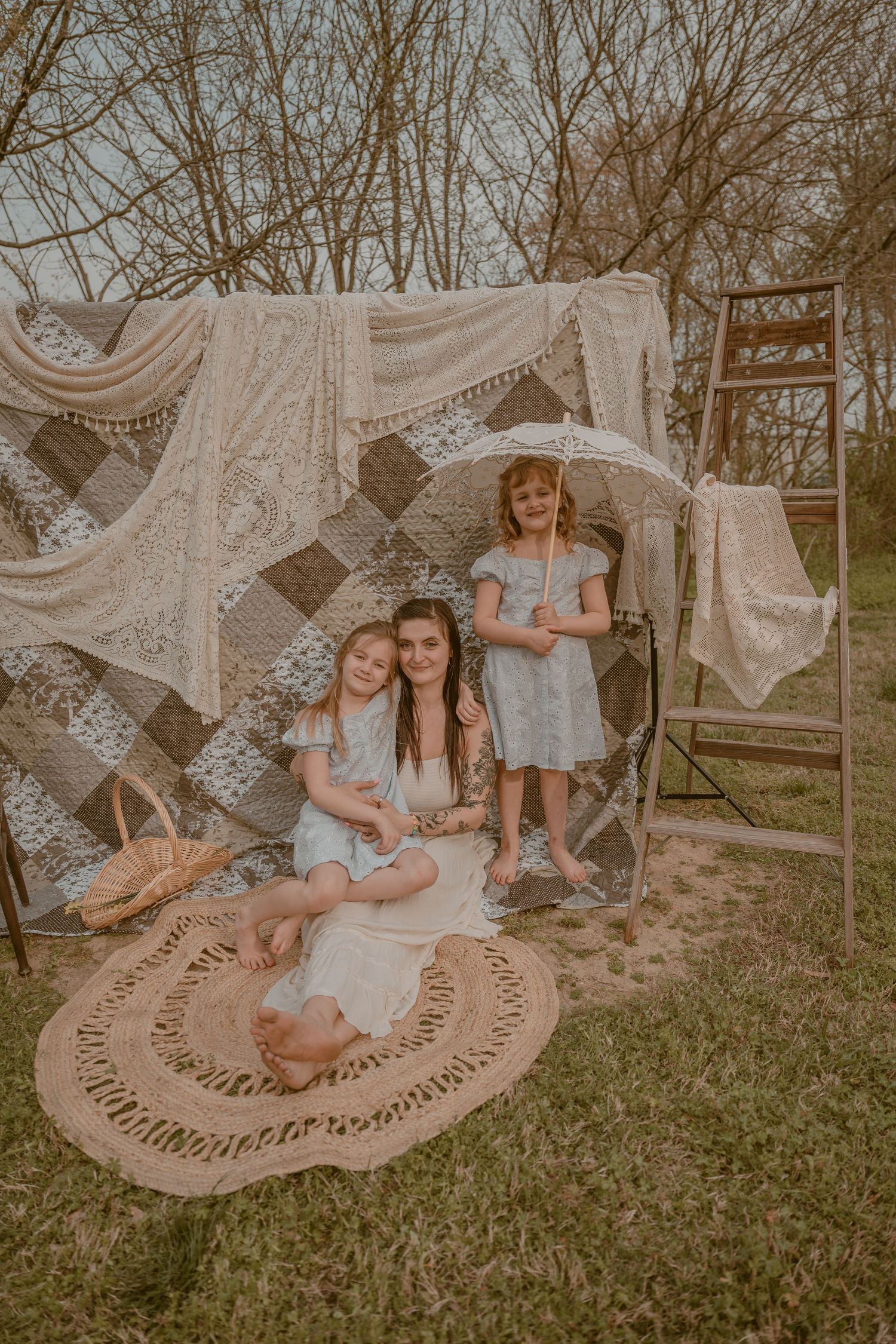 Mother and two daughters pose outdoors under a boho-style canopy of quilts and lace on a woven rug.