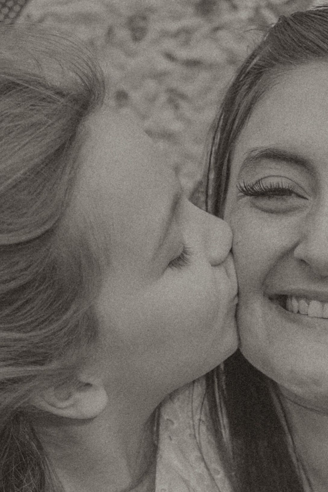 A young girl kisses the cheek of a smiling woman in a tender black and white portrait photo.