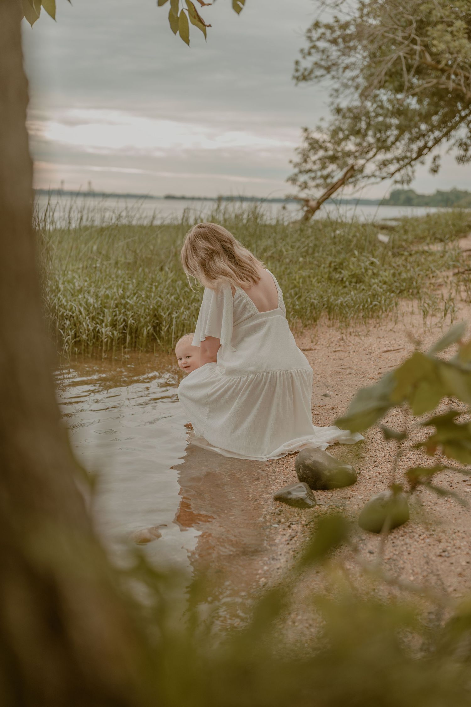 Woman in white dress crouching by water's edge with baby, surrounded by reeds and trees at dusk.